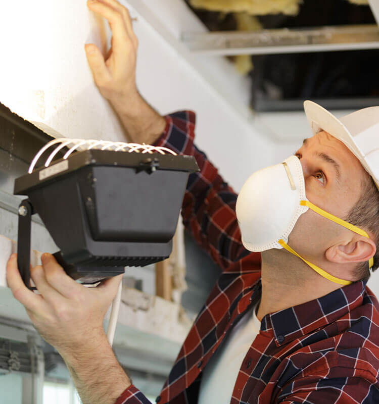 Ein Mann schaut mit Maske, Helm und Baustrahler unter den Dachstuhl